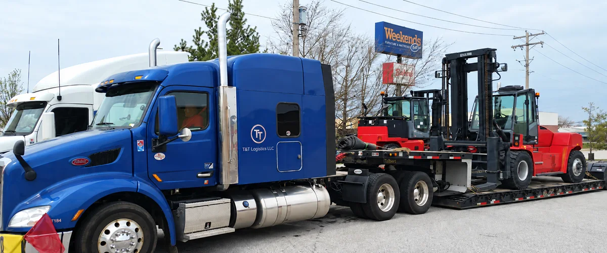 A blue T&T tractor transporting red forklifts on a flatbed trailer
