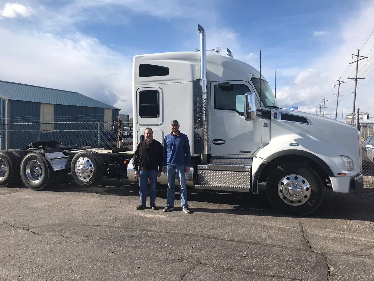 Two people standing beside a white Kenworth tractor in the yard